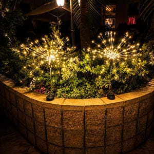 Op de afbeelding: Twee bolvormige solartuinlampen met dunne, stralende metalen stelen en warmwitte led-lampen die een vuurwerkeffect creëren. Elke lamp heeft een kleine, zwarte zonnepaneelvoet die op een stenen muur tussen het groen is geplaatst.