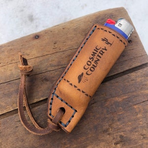 May include: A brown leather lighter case with a blue lighter inside. The case has a stitched design and the text "COSMIC COUNTRY" with a UFO graphic is engraved on the front.