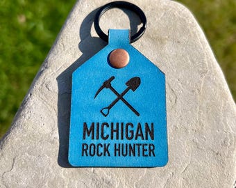 May include: A blue leather keychain with a black key ring. The keychain has a copper rivet and features a crossed pickaxe and shovel design with the words "MICHIGAN ROCK HUNTER" in black.