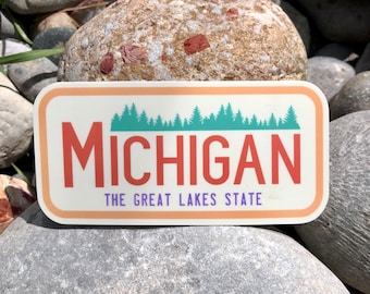 May include: A sticker with a white background and a peach border. The sticker features a teal silhouette of a forest with the word "MICHIGAN" in red text and the words "THE GREAT LAKES STATE" in purple text.