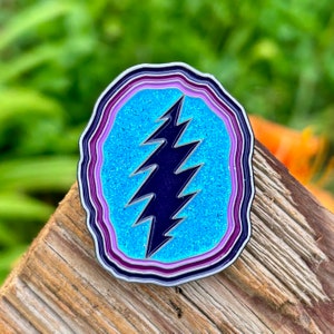 May include: A blue and purple glitter enamel pin with a lightning bolt design. The pin is shaped like a geode with multiple layers of purple and white.