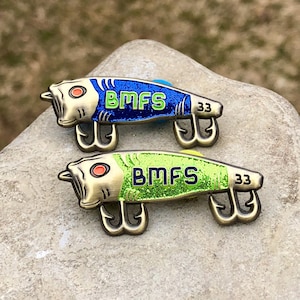 May include: Two fishing lure pins with a metallic finish. One is blue with the letters "BMFS" and the number "33", the other is green with the same text and number. Both have three hooks and red eye details.