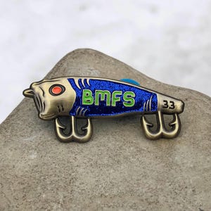 May include: A bronze-colored fishing lure pin with a blue glitter body and the letters "BMFS" in green. The pin has two treble hooks and the number "33". The lure has a red eye and is resting on a gray rock.