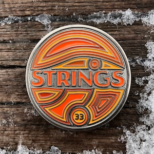 May include: Circular metal pin with the word "STRINGS" in orange lettering. The pin has an orange and yellow design with the number "33" at the bottom. The pin is set against a wooden background with snow.