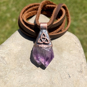 May include: A handcrafted amethyst pendant with a copper-colored metal setting and a brown leather cord. The pendant features a Celtic knot design. The purple crystal is a natural, pointed shape. The leather cord is coiled.