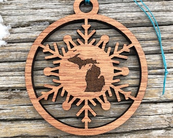 May include: Wooden Christmas ornament featuring a snowflake design with the state of Michigan in the center. The ornament is made of light brown wood and has a blue string for hanging. The design is laser cut.