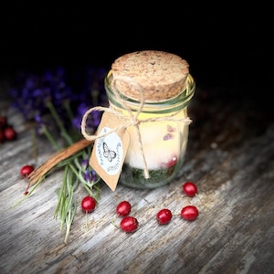 May include: A small glass jar candle with a cork stopper, featuring a light-colored wax interior and a green base layer. A brown paper tag with a black butterfly illustration is tied to the jar with twine, accompanied by purple lavender sprigs and red berries.