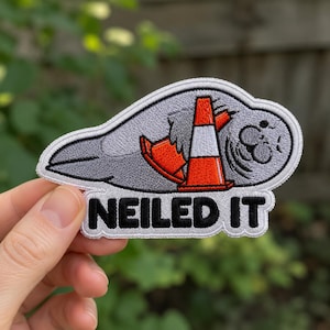 May include: An embroidered patch featuring a gray manatee with a closed-eye expression, partially covered by an orange and white traffic cone. The text "NEILED IT" is stitched in bold black capital letters at the bottom of the patch.