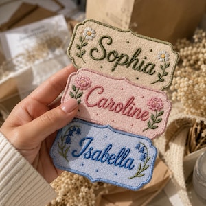 May include: Three rectangular embroidered patches with scalloped edges, featuring the names Sophia, Caroline, and Isabella in cursive script. Each patch displays a unique floral motif and a dotted pattern on a beige, pink, or blue background.