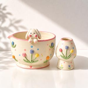 May include: A cream-colored ceramic mixing bowl and a matching small vase, both decorated with raised, colorful tulip motifs and tiny painted flowers. The bowl features a sculpted white bunny figure perched on its rim and a pink painted edge.