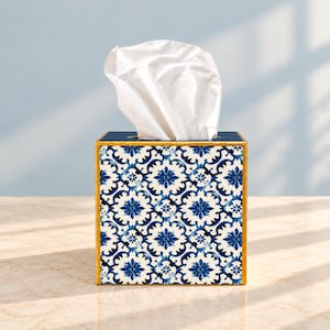 May include: A square tissue box cover featuring a blue and white floral tile pattern with gold-toned edges. The box is filled with white tissues that extend from the top opening, displaying a repeating geometric design on all visible sides.