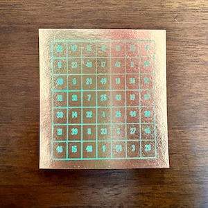 May include: A square metallic copper-colored sticker featuring a grid of seven columns and seven rows. Each cell contains a unique number printed in a teal-colored ink. The grid lines are also outlined in the same teal color against the reflective surface.