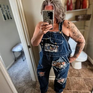 May include: Dark blue denim overalls featuring a distressed, frayed aesthetic with multiple patches sewn onto the chest pocket and legs. The garment includes metal hardware, a front bib pocket, and side pockets, showcasing a worn-in, textured appearance.
