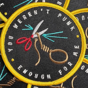 May include: A circular black embroidered patch with a yellow border. The design features a pair of silver scissors with red handles cutting a tan rope tied in a loop. The text "YOU WEREN'T PUNK ENOUGH FOR ME" is stitched in white around the edge.