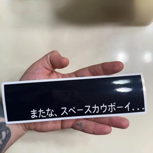 May include: A rectangular sticker with a black background and a thin white border. The sticker features white Japanese text that reads "またな、スペースカウボーイ..." which translates to "See you, space cowboy..." in a clean, sans-serif font.