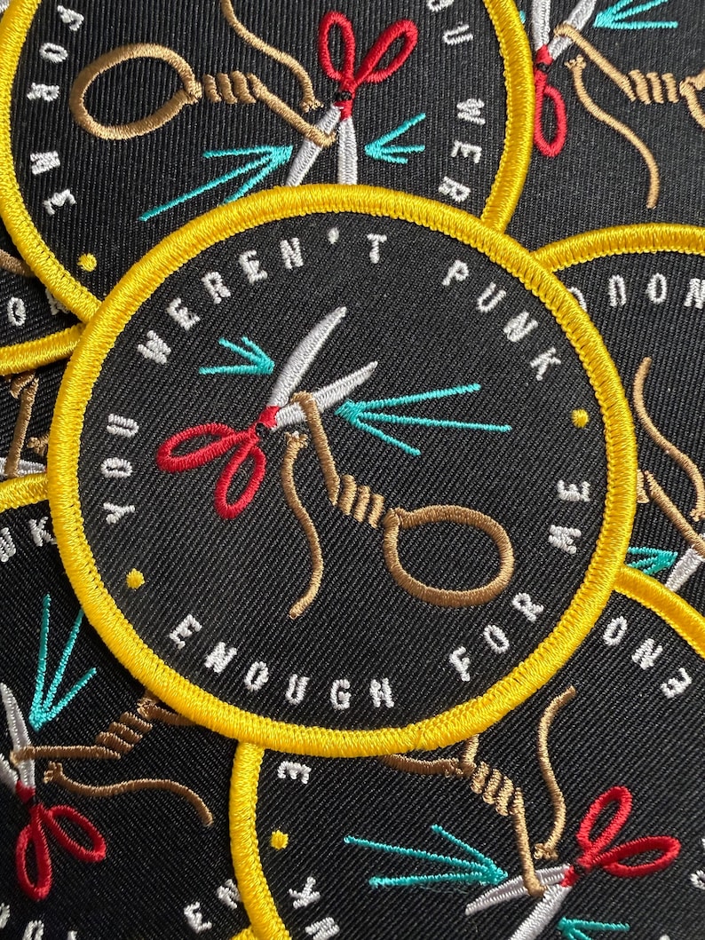 May include: A circular black embroidered patch with a yellow border. The design features a pair of silver scissors with red handles cutting a tan rope tied in a loop. The text "YOU WEREN'T PUNK ENOUGH FOR ME" is stitched in white around the edge.