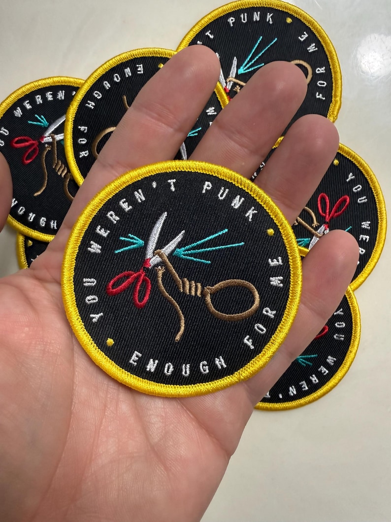 May include: A circular embroidered patch with a black background and a bright yellow border. The design features a pair of scissors with red handles and a brown rope loop. The text "YOU WEREN'T PUNK ENOUGH FOR ME" is stitched in white around the edge.
