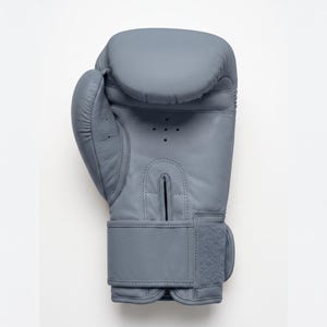 May include: A solid slate gray boxing glove featuring a smooth exterior, a padded knuckle area, and a thumb section. The palm side includes a vertical ventilation slit with four small circular perforations above it and a wide adjustable wrist strap.