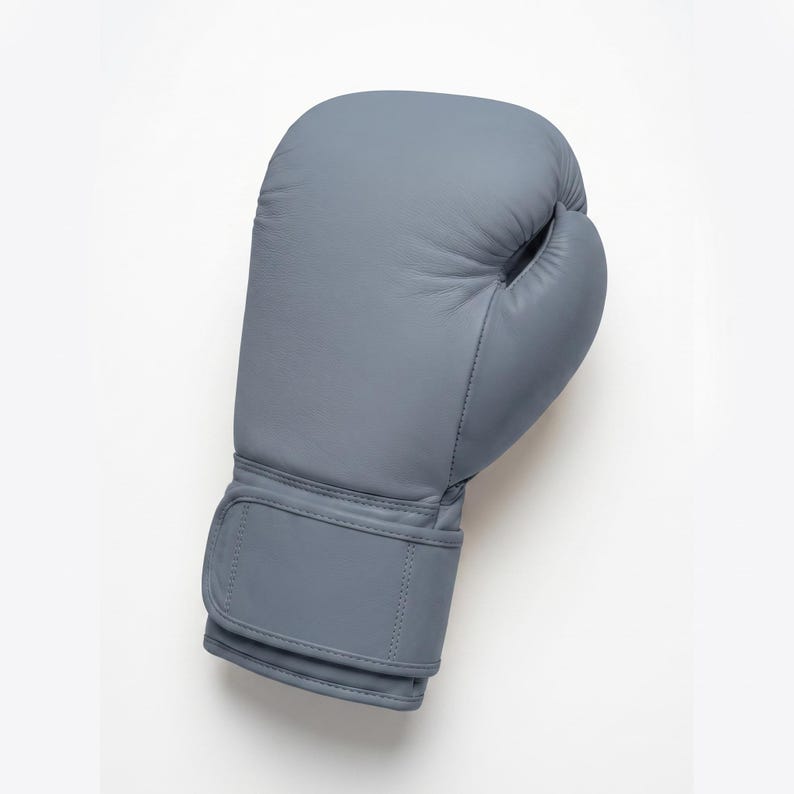 May include: A solid slate gray boxing glove featuring a smooth, matte finish, a padded knuckle area, and a wide wrist strap with a hook-and-loop closure system. The glove is constructed with visible stitching along the seams and a thumb section.