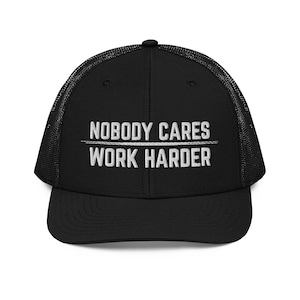May include: A black trucker hat featuring a mesh back and a solid front panel. The front displays white embroidered text that reads NOBODY CARES followed by a horizontal line and the text WORK HARDER. The cap has a curved brim and a button top.