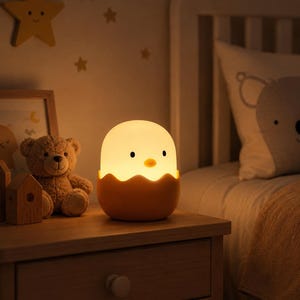 May include: A glowing, egg-shaped night light featuring a smooth, off-white top and a yellow, cracked-shell base. The lamp has two small black eyes and a tiny orange beak, emitting a soft, warm light that illuminates the surrounding area.