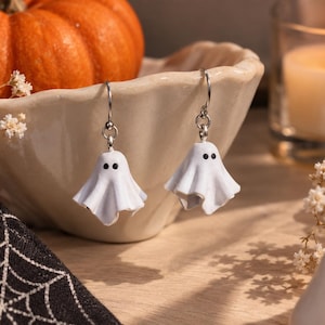 Peut inclure: Une paire de boucles d'oreilles pendantes avec de petites breloques blanches en forme de fantôme, dotées de deux points noirs pour les yeux. Les fantômes sont suspendus à des crochets en métal argenté, formant un design simple et net.