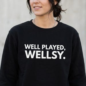 May include: A black crewneck sweatshirt featuring the white text "WELL PLAYED, WELLSY." printed in a bold, sans-serif font across the chest. The garment has long sleeves and a ribbed neckline, providing a simple and clean design for casual wear.