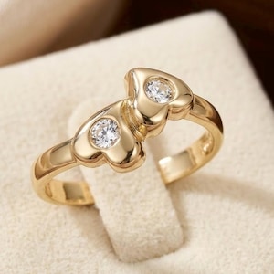 May include: A gold ring featuring two heart-shaped settings, each containing a single round clear gemstone. The band is smooth and polished, with the two hearts positioned side-by-side at the center of the ring, creating a continuous design.