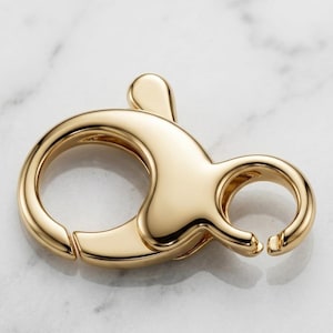 May include: A polished gold-tone lobster claw clasp featuring a smooth, rounded metal surface with a spring-loaded mechanism. The jewelry finding has a circular loop on one side and a curved, hinged opening on the other for secure attachment.