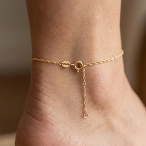 May include: A gold-toned rope chain anklet featuring a circular spring ring clasp and a short dangling extension chain. The delicate twisted metal links form a continuous band that secures around the ankle with a small metal connector.
