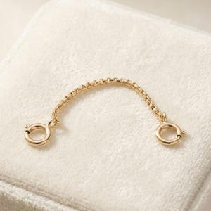 May include: A gold-toned chain extender featuring a delicate box link design. The chain is curved in a semi-circular shape and is finished with a circular spring ring clasp at each end, providing a secure connection for jewelry pieces.