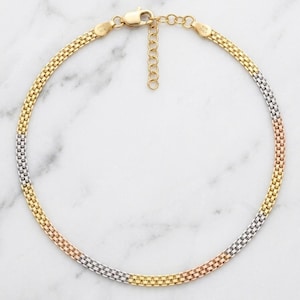 May include: A tri-color chain necklace featuring a woven, square-link pattern in alternating sections of yellow, rose, and white gold tones. The necklace includes a lobster claw clasp and an adjustable extender chain with small circular links.