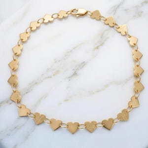 May include: A gold-toned chain necklace composed of a series of repeating heart-shaped links connected by small circular jump rings. The necklace features a lobster claw clasp closure at the top, with each heart link displaying a smooth, polished finish.