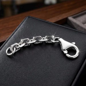 May include: A polished silver-tone chain extender featuring a series of rectangular links and a large, secure lobster claw clasp. The metal surface has a high-shine finish that reflects light, highlighting the smooth, rounded edges of each link.