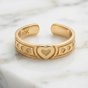 May include: A gold open-band ring featuring a repeating heart pattern along the outer surface. The interior of the band is clearly stamped with the text 10K, indicating the gold purity. The design consists of a central heart flanked by smaller heart motifs.