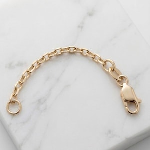 May include: A gold-toned metal chain extender featuring a series of uniform oval links, a small circular jump ring at one end, and a secure lobster claw clasp at the other end, designed to increase the length of a necklace or bracelet.