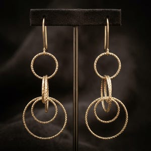 May include: A pair of gold-toned drop earrings featuring three interlocking circular rings of varying sizes. Each ring has a textured, shimmering surface. The earrings are suspended from thin, curved ear wires that attach to the top ring.