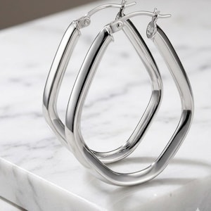 May include: A pair of polished silver hoop earrings with a distinct teardrop or pear-shaped silhouette. These earrings feature a smooth, reflective metallic surface and a secure hinged post closure mechanism at the top for easy wear and removal.