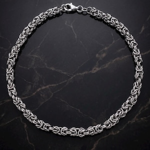 May include: A silver-toned chain necklace featuring a complex Byzantine weave pattern. The necklace is constructed from numerous interlocking metal rings and is secured with a polished lobster claw clasp at the top center of the circular arrangement.
