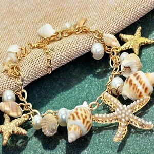 May include: A gold-tone chain bracelet featuring an assortment of white pearls, various small tan and brown patterned seashells, and two gold-tone starfish charms. The bracelet is constructed with a delicate metal link chain connecting each element.