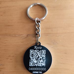 May include: A round black metal pet tag attached to a silver split ring and chain. The tag features the name Rosie in white script, a white QR code, the number 0400000000, and the text ZOOMETAG printed in white on the front surface.