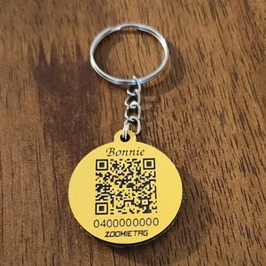 May include: A round yellow metal pet tag attached to a silver split ring. The tag features the name Bonnie in script, a black QR code, the number 0400000000, and the text ZOOMIETAG printed in black on the front surface of the circular metal disc.