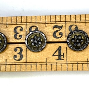 Puede incluir: Cuatro botones antiguos de plata y oro con un diseño de flor. Los botones están alineados en una regla de madera con los números 3, 4 y 5 visibles.