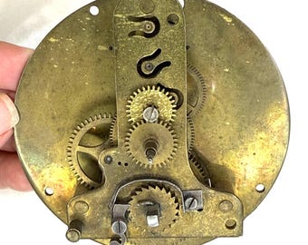 May include: A detailed view of a brass clock mechanism. It displays numerous gears, a central vertical plate, and a circular base. The brass has an aged look, with signs of wear and discolouration. The mechanism is likely from a vintage clock.