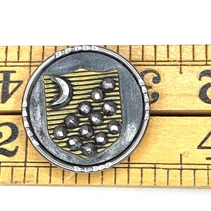 Peut inclure: Un bouton en argent et or avec un croissant de lune et un motif de bouclier. Le bouclier a un fond doré avec un motif de lignes et un groupe de petites pierres rondes noires.