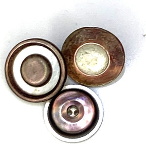 Puede incluir: Tres botones redondos de metal con diferentes diseños. El primer botón es un botón marrón con un anillo blanco alrededor del centro. El segundo botón es un botón marrón con un centro blanco. El tercer botón es un botón morado con un centro plateado.