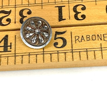 May include: A silver and gold button with a raised, geometric flower design. The button is about 1 inch in diameter and is sitting on a wooden ruler.