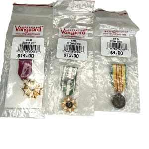 May include: Three military medals in plastic packaging. The medals include a Legion of Merit, a RW Champion, and a Vietnam Service medal. Each medal has a ribbon and is gold-colored with intricate details.