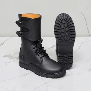 Puede incluir: Una bota negra de media caña con cordones en la parte delantera y una solapa ancha asegurada por dos hebillas laterales. La bota tiene una puntera redondeada, una suela gruesa con tacos y un forro interior color canela. Se muestra otra bota desde atrás.