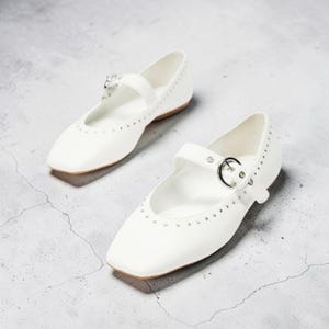 May include: A pair of white flat shoes featuring a square toe design, a single strap across the instep with a silver-toned metal buckle, and a row of small silver-toned studs lining the opening of the shoe. The shoes have a low profile.